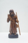 Escultura "O Frade Viajante" em Madeira Assinada Mestre Dunga.Esta singular escultura em madeira maciça é uma obra autêntica de Arte Popular Brasileira do renomado Mestre Dunga, com sua assinatura "DUNGA" entalhada na base; a peça retrata de forma expressiva um Frade ou Peregrino, envolto em um hábito com capuz, que não porta símbolos hagiográficos tradicionais; a figura sustenta um cajado em uma mão e um cantil ou garrafa sobre o ombro, sugerindo a jornada e a provisão do viajante; o entalhe é vigoroso, característico da arte regional do Nordeste, e possui acabamento em verniz escuro; é uma peça de forte valor cultural que celebra a iconografia da vida no sertão. Assinado no fundo. Alt. 31 cm.
