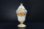 Antiga compoteira em faiança portuguesa, adornada com padrões florais em tons de laranja e amarelo, com folhagens e frisos em verde e marrom. Base em forma de pedestal, corpo bojudo e uma tampa cônica. Apresenta uma textura em relevo no padrão escamas. Alt. 29 cm. .