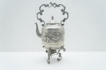 Magnífico Samovar (Chaleira em Suporte), metal prateado. Datado dos meados do Século XX, é uma peça de colecionador com suntuosa decoração em baixo-relevo de cenas figurativas e florais no corpo, complementada por alça alta de arabescos. Um ícone de luxo e hospitalidade. Alt. 40 cm.