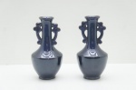 par de vasos decorativos em porcelana oriental, destacando-se pela sua forma elegante e um esmalte de acabamento profundamente sofisticado.Cada vaso mede 18 cm de altura e possui um corpo bojudo que se afina em um gargalo estreito. O ponto focal da peça reside no seu esmalte único: uma camada de cor marrom-escura (próxima ao berinjela ou preto) com um fundo azulado intenso. Sob a luz, o esmalte revela um efeito deslumbrante, iridescente e perolado, que remete à técnica clássica chinesa Oil Spot (ou "Mancha de Óleo"), conferindo um brilho vítreo e profundo à superfície.O design é enriquecido pelas alças moldadas no gargalo, trabalhadas em forma de motivos arcaicos ou dragões estilizados. Estas alças apresentam um esmalte em azul-cobalto vibrante, criando um contraste visual impactante com o corpo escuro. O acabamento é altamente lustroso, realçando a complexidade e a variação da cor do esmalte.