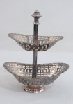 Fruteira com dois estágios em metal espessurado a prata, base redonda, duas sessões vazadas decoradas em geométricos e gotas, haste finalizada com pega em formato de pinha. Marcada TB. Med. 20 x 17 x 12 cm.