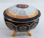 Caixa porta joias oval em fina porcelana alemã esmaltada na tonalidade azul cobalto, ornada com cena angelical com farta douração, base tripóide. Marcada. Med. 9 x 10 cm diâmetro. Parte interna da tampa com discreto bicado.