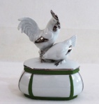 Curioso pimenteiro oval em porcelana branca com detalhes em verde e prateado, Tampa encimada com casal de galináceos com detalhes prateados. Marcada na base em relevo. Med. 15 x 11 x 7,5 cm.