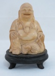 Escultura de buda em material natural na cor marfim, apoiado sobre peanha de madeira. Med. 14 x 9 x 8 cm.