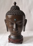 Escultura em bronze representando "Cabeça de Buda" com inserção de pedra azul na testa. Tibete. A cabeça é uma das partes do corpo mais importantes do Buda, tendo vários simbolismos, como a expressão de serenidade que demonstra estar em posição de meditação e as orelhas mais compridas que representam estar ouvindo o outro. Base de peanha em madeira. Peso 4.800 gramas. Med. 42 cm alt x 23 cm diâmetro.