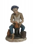 Escultura em porcelana pintada à mão, representando oleiro no torno de cerâmica, ricamente detalhada. Base forrada em veludo vermelho, com etiqueta Made in Taiwan. Mede 18cm x 13cm.