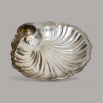 petisqueira ou centro de mesa em metal prateado, moldado em formato de concha. É uma peça inglesa, frequentemente associada à manufatura Old English Silver & Co., Sheffield. O design inclui um interior côncavo com frisos e bordas recortadas em arcos. Possui um receptáculo específico para molho ou manteiga, como visto no canto superior esquerdo. Essas peças são geralmente feitas de metal espessurado a prata (banho de prata de alta qualidade). Medindo 28 de larg x 29 comp