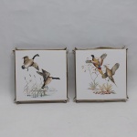 Dois suportes para pratos quentes, manufaturados a partir de azulejos alemães decorados com patos e faisões. Guarnição em metal dourado. Med. 17 x 17 cm.