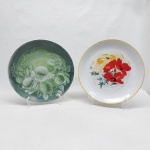 Dois pratos para servir em porcelana: A) Branco, borda amarela. Flores, folhagens e ramos de trigo ao centro. Diam. 26 cm. B) Verde, decorado com flores e folhagens pintadas a mão. Diam. 27 cm.