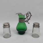 Três peças para galheteiro em vidro verde, translúcido e metal prateado: A) Recipiente p/azeite em vidro verde, tampa aplicada com cacho de uvas, guarnição vasadas com flores e alça na forma de acantos. Alt. 11 cm. B) Saleiro e pimenteiro translúcido, tampa metálica. Alt. 7 cm.