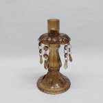 Gracioso castiçal em vidro na tonalidade âmbar, estilo europeu. Profusamente lapidado com caneluras e motivos. Bobèche ao gosto bico de jaca, 4 argolas com pingentes (falta uma). Alt. 21 cm.
