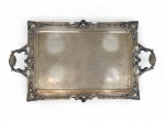 PRATA DE LEI - Grande bandeja / tabuleiro em prata de lei. Elegante bandeja retangular em prata de lei, com rica decoração gravada em volutas e arabescos de inspiração rococó. Bordas recortadas com moldura em relevo e alças vazadas ornamentadas por volutas e florões, típicas do trabalho de prateiros ingleses e brasileiros da primeira metade do século XX. Peça de grande presença, ideal para centro de mesa, bar ou serviço de chá. Medidas: 64 cm (comprimento total, incluindo alças) x 42,5 cm (largura). Peso: 5.592 gramas Prata de lei. Procedência: Acervo família Rezende.* ATENÇÃO. Foto complementar apenas a título de ilustração. Este lote refere-se UNICAMENTE a bandeja de prata não incluindo demais itens.