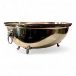 METAL PRATEADO - Grande recipiente / cachepô (centro de mesa) em metal espessurado a prata.Peça de grandes dimensões, formato oval, decorada com cabeças de leão nas laterais, cada uma portando argola móvel. Borda superior com friso torcido e quatro pés em volutas e folhagens, em estilo neoclássico tardio. Interior com intenso desgaste do banho e exterior com marcas leves de uso sobretudo na borda.Dimensões: 43 cm de comprimento x 34 cm de largura x 17 cm de altura.Peça imponente e versátil  pode ser utilizada como centro de mesa, cachepô para flores ou champanheira individual. Faz par com o lote seguinte.