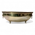 METAL PRATEADO - Grande recipiente / cachepô (centro de mesa) em metal espessurado a prata.Peça de grandes dimensões, formato oval, decorada com cabeças de leão nas laterais, cada uma portando argola móvel. Borda superior com friso torcido e quatro pés em volutas e folhagens, em estilo neoclássico tardio. Interior com intenso desgaste do banho e exterior com marcas leves de uso e pontos de oxidação discreta, sobretudo na borda.Dimensões: 43 cm de comprimento x 34 cm de largura x 17 cm de altura.Peça imponente e versátil  pode ser utilizada como centro de mesa, cachepô para flores ou champanheira individual. Faz par com o lote anterior.