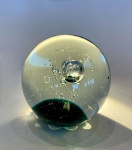Peso de papel em vidro artístico, com inclusão de bolhas e base verde-esmeralda. Europa, meados do século XX.Grande peso de papel em vidro soprado maciço, formato esférico, com interior composto por massa verde translúcida e núcleo de bolhas de ar aprisionadas, formando desenho tridimensional. Trabalho artesanal de grande pureza técnica, com transparência cristalina e leve iridescência superficial.Etiqueta sob a base com marca não identificada, parcialmente legível (dor), possivelmente vinculada a oficinas escandinavas ou italianas de vidro artístico (Murano-style).Apresenta apenas ínfima bolha de ar na superfície, sem lascas nem riscos.Diâmetro: 10 cm.Nota técnica:A técnica de inclusão de bolhas (controlled bubbles ou inclusions à bulles) é típica do vidro artístico europeu do pós-guerra, especialmente nas produções de Murano (Itália), Kosta Boda (Suécia) e Caithness (Escócia). Peças dessa natureza, popularizadas como elementos de decoração moderna, foram também exportadas para a América e o Brasil nas décadas de 196070.