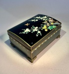Caixa de música japonesa com decoração em esmalte cloisonné e paisagem dourada interna. Fabricante Sankyo. Japão, meados do século XX.Caixa musical retangular em metal esmaltado e dourado, decorada na tampa com ramagens floridas e pequenos colibris em voo sobre fundo negro  composição típica do cloisonné japonês de meados do século XX, de grande delicadeza e precisão técnica.Interior com douração integral e gravações em baixo-relevo representando o Monte Fuji, barcos de pesca e paisagem lacustre.Mecanismo musical da marca Sankyo  Japan, em perfeito funcionamento, emitindo melodia tradicional japonesa de época.Medidas: 14 cm x 9,5 cm x 6,5 cmApresenta leve trinca no esmalte lateral, sem perda de estabilidade. Marcas naturais do tempo.Nota histórica:A Sankyo Seiki Co., fundada em 1946 em Tóquio, tornou-se o principal fabricante japonês de mecanismos musicais de corda, exportando amplamente durante as décadas de 1950 a 1970.Caixas como esta, ricamente decoradas com esmalte e paisagens orientais douradas, foram produzidas como objetos de luxo e lembranças para exportação, simbolizando o refinamento do artesanato nipônico pós-guerra.