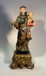 Santo Antônio com Menino Jesus  manufatura Velloso & Irmão, Lisboa. Portugal, século XX.Imagem devocional em madeira entalhada e policromada, representando Santo Antônio de Pádua segurando o Menino Jesus, ambos com fisionomias serenas e vestes ornamentadas. O hábito franciscano apresenta ricamente pintados motivos dourados sobre fundo escuro, com estofamento a ouro e pintura em padrões estilizados. O conjunto repousa sobre base quadrada com talha dourada e elementos fitomórficos.Na parte inferior, conserva o selo original da manufatura Velloso & Irmão  Santeiro com Ornamentos Religiosos  Lisboa, Rua do Crucifixo, 90, importante casa lisboeta especializada em imaginária sacra e objetos litúrgicos, ativa entre o fim do século XIX e meados do XX.Medidas: 33 cm de altura x 13 x 13 cm (base).Estado de conservação: desgaste compatível com a idade; pequenas perdas na policromia e douração, sem comprometimento da integridade estrutural.