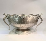 L.S. Gaillard, Paris (c. 19001930)Grande centerpiece ou cachepot em metal prateado, corpo canelado e bordas recortadas com ornamentação rocaille e volutas florais.Alças laterais em forma de grifos alados, ricamente fundidas.Selo de manufatura em relevo LSG sob grifo e número de modelo 8506.Medidas: 36 cm de diâmetro x 22 cm de altura.Peça de exceção, de proporções monumentais, representativa do gosto francês da Belle Époque, período de apogeu da ourivesaria decorativa. A elegância das curvas e a riqueza dos relevos atestam produção industrial de alto padrão, voltada à exportação.Nota do editor: A manufatura L.S. Gaillard, empresa francesa de metal prateado esteve ativa entre o fim do século XIX e as primeiras décadas do XX, sendo estabelecida em Paris (rue des Archives) e especializada em orfèvrerie galvanizada e peças de mesa de estilo Luís XV e Luís XVI. O grifo (ou águia estilizada) foi usado por Gaillard como selo de qualidade em ligas prateadas, e o número 8506 indica o modelo de referência no catálogo da fábrica, o que confirma série de produção. Peça faz par com o lote anterior. Imponente e completa.