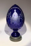 Pinha em cristal azul-cobalto lapidado e gravado com igreja ortodoxaEuropa Central ou Rússia, meados do século XXCristal de chumbo em tom azul-cobalto profundo, de alta densidade e pureza ótica, lapidado e gravado em relevo polido com representação de uma igreja ortodoxa russa de cúpulas bulbosas, ladeada por árvores e ornamentações vegetais. O verso exibe lapidação em cruz, composta por círculos facetados e elementos em relevo que se iluminam sob incidência de luz, criando efeito translúcido notável. Apoiada sobre base de bronze patinado, de tom verde-azulado.Altura: 20 cmDiâmetro máximo: 9,5 cmO azul intenso é obtido pela adição de óxidos de cobalto ao vidro em fusão, processo que confere cor homogênea e profundidade incomum. A gravação, executada em múltiplos planos e com acabamento polido em zonas selecionadas, evidencia técnica refinada típica dos grandes ateliês boêmios e russos do pós-guerra, voltados à produção de objetos de devoção e prestígio.Nota iconográfica curta. A igreja ortodoxa gravada simboliza o coração espiritual do mundo eslavo  a casa de oração como microcosmo da fé cristã oriental. A lapidação em cruz no verso reforça o caráter devocional da peça, aludindo à interseção entre o misticismo bizantino e a arte do vidro centro-europeia. O azul-cobalto, cor do infinito e da transcendência, amplifica o simbolismo da eternidade e da comunhão entre o divino e o terreno.Este lote faz par com o lote anterior.