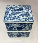 Caixas sobrepostas  Porcelana chinesa azul e brancaConjunto formado por duas pequenas caixas quadradas com tampa, em porcelana de fina espessura e esmalte translúcido.Decoração em azul-cobalto sob o vidrado, representando ramos floridos e pequenos pássaros (colibris) entre folhagens estilizadas, num desenho contínuo que envolve todas as faces da peça.Selo aposto sob o esmalte na base, de formato quadrado.Medidas do conjunto: 7 cm de altura x 6 cm de base.Leitura: exemplar moderno de tradição oriental, reinterpretando a estética Qing tardia dos blue & white wares, com leveza de traço e composição de caráter lírico  o voo dos pássaros simboliza prosperidade e harmonia doméstica.