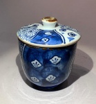 ARTE ORIENTAL - Pequeno recipiente com tampa  Japão, século XX. Cerâmica oriental de corpo fino e paredes verticais, com tampa dotada de pega central e pequeno orifício para saída de vapor.Decoração sob o vidrado em azul-cobalto (sometsuke), com reservas alternadas em padrões geométricos e nuvens estilizadas, representando o contraste entre o céu diurno e o noturno.Assinatura do artesão no fundo.Medidas: 10 cm de altura x 7,5 cm de diâmetro.Exemplar de chawan com tampa (mushimono chawan), utilizado tradicionalmente para servir chá quente, caldos leves ou preparos cozidos no vapor, típicos da culinária japonesa refinada.O pequeno orifício na tampa permite a liberação do vapor e evita a condensação sobre o alimento.A borda apresenta efeito ferruginoso intencional, conhecido como renga-yaki, evocando o aspecto terroso e ancestral das cerâmicas de forno de pedra.Nota cultural: Durante o kaiseki - refeição ritual que precede a cerimônia do chá - cada recipiente é escolhido em harmonia com a estação, o tema poético e a textura da comida.O mushimono chawan é símbolo de acolhimento e delicadeza, expressando a filosofia japonesa do ichigo ichie (cada encontro é único), em que o gesto de servir algo quente traduz respeito e presença espiritual.