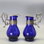 DEMI CRISTAL - Antigo par de galheteiro em tom azul, com acabamento em metal espessurado a prata, tampa em relevo com cachos de uvas. Alt. 12 cm. Marcas do tempo e de uso.