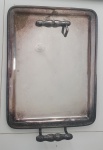 Bandeja retangular de metal com acabamento prateado ou cromado, apresentando sinais de uso e manchas. Possui bordas elevadas e duas alças ornamentadas em estilo clássico nas extremidades curtas. A superfície central é lisa, polida e refletora. - Medidas: 36x46 cm Lote CPM alças solta.