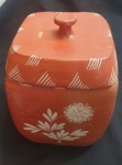 Pote de cerâmica artesanal com tampa, de formato retangular e cantos suavemente arredondados. Possui pintura em tom terroso avermelhado e delicados adornos brancos feitos à mão: na parte frontal, destaca-se um ramo com folhas e uma flor estilizada; já na tampa, padrões diagonais se repetem ao longo das bordas. O puxador, central, é modelado em forma de botão arredondado, conferindo praticidade e charme. A peça apresenta estilo rústico, com possível inspiração na arte popular indígena.Medidas: 19 x 19 x 29 cm. - Mote com marcas de uso, alguns lascados.