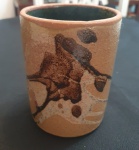 Vaso cilíndrico em cerâmica, de acabamento rústico e tonalidade terrosa. Sua superfície apresenta decoração abstrata em tons escuros, próximos ao marrom e preto, com efeitos que lembram manchas orgânicas ou padrões naturais formados durante o processo de queima ou aplicação do esmalte. parece a técnica japonesa chamada Neriage - Altura: 13 cm e Diâmetro: 10 cm
