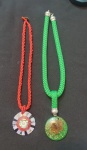 Dois cordões artesanais: um em contas vermelhas com pingente circular colorido em mosaico e espiral central, de estilo vibrante; outro em fio trançado verde com pingente em resina translúcida, dourados e flor seca, de inspiração natural e orgânica.