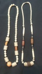 Dois cordões artesanais de estilo rústico, confeccionados com materiais diversos e detalhes geométricos. Um é composto por contas esféricas claras, intercaladas por discos achatados e peças em madeira, destacando-se pelo grande bead arredondado em tom marrom mesclado na parte inferior. Já o outro traz contas menores em tom claro, combinadas com cilindros alongados, madeira escura e detalhes metálicos, conferindo-lhe um visual mais alongado e tribal. Ambos refletem um design étnico e artesanal, de forte apelo decorativo e autêntico.