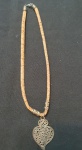 Cordão artesanal comprado em Portugal, feito com contas cilíndricas em tom bege claro, intercaladas por detalhes metálicos. O pingente é uma peça prateada em formato de coração estilizado, trabalhado em filigrana, que remete à tradição portuguesa. Uma peça rústica e elegante, de inspiração cultural.
