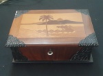 Caixa de joias antiga/vintage em madeira, com detalhes em embutidos e cantoneiras metálicas decorativas. A tampa exibe um refinado trabalho de marchetaria, formando uma paisagem tropical com palmeiras e montanhas. Os cantos são ornamentados com ferragens em metal ricamente trabalhadas em formato de folhas.- Medidas: 25x18x9 cm