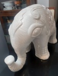 Escultura de elefante de porcelana branca com detalhes em relevo, que parecem ser esculpidos diretamente na superfície. O elefante tem um acabamento vitrificado, proporcionando um brilho suave que reflete a luz. Suas características, como os olhos e as orelhas, estão bem definidas com texturas onduladas e padrões simétricos que cobrem boa parte do corpo. A tromba é levantada, uma posição tradicionalmente associada a boa sorte. As patas do elefante são curtas e robustas, e o corpo possui uma postura sólida e estável. A peça é decorativa e possui um ar elegante, destacando-se pela simplicidade da cor e pela riqueza dos detalhes esculpidos. LOTE COM RACHADO NA TROMBA E COM PEQUENO LASCADOS. Medidas:36x17x16 cm