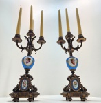 Par de candelabros em porcelana Sèvres e bronze dourado, França, séc. XIX Magnífico par de candelabros franceses em porcelana Sèvres, no clássico tom bleu céleste, com montagens em bronze dourado ricamente cinzelado. Cada peça apresenta corpo em porcelana com medalhões pintados à mão, representando delicados retratos femininos ao gosto do século XVIII, emoldurados por guirlandas e arabescos dourados. A base, igualmente adornada, ostenta cartelas ovais florais em esmalte policromado, e sustenta estrutura de quatro braços sinuosos para velas, com minucioso trabalho de fundição e acabamento. Dimensões: 45 x 17 centímetros sem velas.