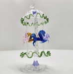Carrossel em vidro artístico Murano Delicada escultura em vidro artístico de Murano, representando um carrossel com cavalos coloridos, executada com notável precisão artesanal. A estrutura translúcida é adornada por bordas onduladas em verde, sustentando delicadas figuras de cavalos em tons de âmbar, lilás e azul, modeladas à mão com transparência e movimento fluido. A composição expressa a leveza e o encanto do vidro soprado veneziano, evocando o universo lúdico e nostálgico dos antigos carrosséis. Altura: 15x7 centímetros.
