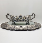 Centro de mesa em prata portuguesa 833  trabalho de ourivesaria barroca. Imponente centro de mesa em prata portuguesa de lei (833 milésimos), composto por base espelhada de moldura profusamente cinzelada e corpo central vazado, com alças laterais em volutas e pés ricamente ornamentados. A decoração apresenta elaborado repertório de inspiração barroca, com conchas, volutas e motivos vegetalistas em composição simétrica, revelando o requinte e o virtuosismo técnico característicos da ourivesaria portuguesa de finais do século XIX e início do XX.O conjunto é marcado pela riqueza do cinzelado e pelo equilíbrio entre o volume escultórico e a leveza do trabalho vazado, resultando em uma peça de forte presença decorativa e excepcional execução artesanal. Apresenta punção da águia portuguesa, correspondente à prata 833, e contraste adicional de oficina, assegurando sua autenticidade e origem nacional. Dimensões: 20 x 70 x 54 centímetros, Peso total: 7.393 kilos.