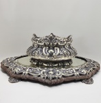 Suntuosa floreira em prata brasileira de lei (900 milésimos), de corpo bojudo e exuberante decoração em relevo, com motivos de conchas, volutas e folhagens, dispostos em composição simétrica de inspiração barroca e rocaille. A peça repousa sobre base moldurada com espelho original, ricamente cinzelada e apoiada em pés ornados, formando um conjunto de notável presença decorativa e refinamento técnico. A execução revela a influência direta da ourivesaria portuguesa e francesa do século XIX, reinterpretada com maestria nas oficinas brasileiras da primeira metade do século XX. Apresenta punção PRATA 900, assegurando a autenticidade do metal e a elevada pureza da liga utilizada. Dimensões: 22 x 64 x 32 centímetros. Peso:6.457 kilos .