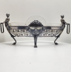WMF  Floreira em metal prateado com taça de cristal gravado  Alemanha, c. 1900Elegante e escultórica floreira da manufatura WMF (Württembergische Metallwarenfabrik), executada em metal prateado (marcação I/O), de corpo oval ricamente vazado e cinzelado com ornamentação de parreiras, guirlandas e mascarões clássicos. Apresenta nas extremidades figuras femininas em pé, em estilo neoclássico, sustentando argolas laterais  composição de refinado equilíbrio entre escultura e funcionalidade, típica das criações da WMF no período Jugendstil / Historicismo tardio. O conjunto é completado por taça interna em cristal gravado, com friso grego e borda recortada, original da manufatura. Marcada na base com o punção WMF e inscrição I/O, utilizados pela casa alemã entre c. 1890 e 1910, assegurando autenticidade e correspondendo à qualidade superior de galvanoplastia. Dimensões: 30 x 64 x 23 centímetros.