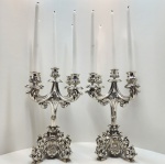 Par de candelabros em prata austro-húngara  Boêmia, c. 1880 a 1910. Marcados com o contraste da cabeça de Diana. Magnífico par de candelabros em prata de lei, cada um com cinco luzes, dispostas em braços sinuosos e profusamente cinzelados, partindo de um fuste central ornamentado. A decoração apresenta exuberantes motivos e folhagens em relevo, alternando volutas, conchas e elementos florais, apoiados sobre bases tripartidas com pés em enrolamentos simétricos, em composição de clara inspiração Rococó. O conjunto reflete o refinamento técnico e o requinte ornamental da ourivesaria austro-húngara do final do século XIX, especialmente das oficinas da Boêmia (atual República Tcheca), conhecidas pela excelência no trabalho em prata e pela fusão entre a tradição barroca e o gosto decorativo centro-europeu. Apresentam o contraste oficial da cabeça de Diana, certificando a origem e autenticidade dentro do sistema de contrastes do Império Austro-Húngaro (1867 a 1922). Dimensões: 40 x 27 centímetros. Peso: 4,506 kilos.