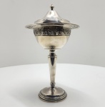 Bombonière de apresentação em prata maciça (Sterling Silver .925), Gorham Silver Co., EUA, década de 1960  modelo nº 7964Elegante bombonière de apresentação em prata maciça (Sterling Silver .925), manufatura Gorham Silver Company, Providence, Rhode Island, Estados Unidos. De proporções esguias e harmonia clássica, apresenta haste elevada e base circular moldurada, sustentando corpo em forma de taça coberta. A borda superior é rendilhada (pierced work), com motivo geométrico refinado, e a tampa ajustada é rematada por pináculo cônico. O desenho alia pureza de linhas e equilíbrio formal, refletindo o estilo neoclássico reinterpretado pela ourivesaria americana do pós-guerra, período em que a Gorham alcançou o auge de sua produção de prata sólida. Marcada sob a base com o símbolo folhado da manufatura, a inscrição STERLING e o número de modelo 7964, indicando prata maciça de 925 milésimos. Dimensões: 34 x 17 gramas. Peso: 620 gramas.