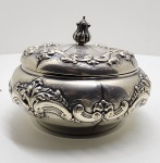 Potiche em prata brasileira (833 milésimos), Ourivesaria União S. Br., São Paulo, c. 1940Imponente potiche com tampa, em prata brasileira de 833 milésimos, manufatura Ourivesaria União, São Paulo, cerca de 1940.De corpo bojudo e tampa ajustada com botão escultórico em forma de fruto estilizado, apresenta ornamentação repuxada e cinzelada em estilo Rococó revivalista, composta por volutas, concheados e motivos florais em relevo, delineados com extremo detalhamento artesanal. A peça exibe o equilíbrio característico da produção paulistana de meados do século XX, fundindo exuberância ornamental e refinamento técnico. Marcada sob a base com os punções UNIÃO S. BR. e PRATA 833, correspondentes à prata nacional de 2º título, amplamente utilizada nas oficinas de São Paulo nas décadas de 1930 e 1950.Dimensões: 13 x 17 centímetros. Peso: 485 gramas.