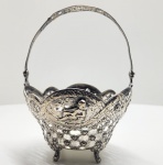 Cestinho em prata vazada e cinzelada com cenas românticas, possivelmente europeia, séc. XIX. Delicado cestinho em prata de formato quadrilobado, sustentado por quatro pés recurvados e alça móvel superior vazada, com ornamentos florais e fitomórficos. As faces exibem painéis ovais em relevo, representando cenas galantes com figuras em paisagem bucólica, enquadradas por molduras de volutas e entrelaçados vazados de extremo detalhamento. A decoração, executada por repujado, cinzelado e recorte, é típica da estética Rococó europeia, inspirada nos motivos franceses do século XVIII  reinterpretada no século XIX por ourives de tradição germânica e inglesa. Dimensões: 18 x 13 centímetros. Peso: 228 gramas.