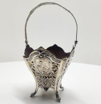 Cestinho em prata portuguesa do início do séc. XX, Delicado cestinho em prata repuxada, cinzelada e vazada, de corpo quadrilobado apoiado sobre quatro pés recurvados e alça móvel superior com decoração rendilhada. As faces exibem painéis ovais repuxados em alto-relevo, representando cenas galantes com figuras em paisagem bucólica, enquadradas por molduras assimétricas de volutas, flores e entrelaçados geométricos  trabalho minucioso de cinzelagem, típico do gosto Rococó português. A peça revela a maestria dos ourives na transposição do repertório decorativo francês setecentista para a prata portuguesa de mesa e ornato. Dimensões: 25 x 14 centímetros. Peso: 437 gramas.