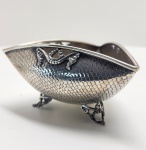 Porta-confeito em prata de formato ovalado, finamente cinzelado em delicado padrão escamado, conferindo textura e luminosidade à peça. A borda superior é realçada por aplicações em relevo com volutas e arabescos simétricos, que acentuam o caráter decorativo e o equilíbrio formal do conjunto. A peça repousa sobre três pés elegantemente rendilhados, de desenho sinuoso e acabamento minucioso, que reforçam a leveza e sofisticação do modelo. O contraste entre as áreas polidas e a pátina escurecida realça a profundidade do trabalho artesanal. Peça de grande refinamento estético, ideal para compor ambientes clássicos ou coleções de prataria artística. Dimensões: 7 x 13 centímetros. Peso: 137 gramas.