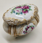 Caixa em porcelana Rosenthal / Bavaria,  Alemanha. Elegante caixa com tampa em porcelana fina alemã, de corpo bojudo e bordas recortadas, apoiada sobre quatro pés curvos. Decoração em flores policromadas  rosas e anêmonas  sobre fundo branco, com detalhes em ouro pintado à mão e finos arabescos dourados em pontilhismo. Tampa com composição floral central e frisos em relevo com douração intensa. Marca em verde sob a base: Rosenthal / Bavaria, com inscrição manuscrita, correspondente à linha decorativa. Dimensões:15x18x13 centímetros.