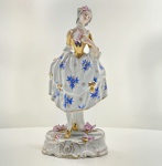 Figura feminina em porcelana Vista Alegre, Portugal, meados do século XX. Figura de dama oitocentista, em porcelana portuguesa moldada e pintada à mão, representando jovem segurando leque. Traje decorado com flores azuis e douradas, corselete em ouro brilhante, e base rococó com pequenas flores aplicadas em relevo. Marca em verde sob a base: V.A. Portugal (Vista Alegre). Dimensões:26 x 11 x 10 centímetros. Estado de conservação: apresenta reparo visível no pescoço, pequenas perdas nas flores aplicadas e leve desgaste do dourado, compatíveis com a idade.