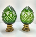 Extraordinário par de pinhas decorativas em cristal de altíssima qualidade, lapidadas e montadas sobre bases em metal maciço e ricamente fundido, de proporções equilibradas e acabamento dourado envelhecido. As pinhas apresentam corpo em cristal duplo verde sobre incolor (overlay), técnica tradicional das cristalerias da Boêmia, obtida pela fusão de duas camadas de vidro soprado e subsequente lapidação profunda à mão, que revela o brilho translúcido do fundo incolor sob o véu verde esmeralda. A lapidação, de traço preciso e simetria exemplar, exibe motivos em losangos, estrelas e cortes em ponta de diamante, típicos da melhor tradição boêmia, reconhecida mundialmente por sua pureza, peso e clareza óptica. O cristal espesso reflete a luz em prismas vivos, produzindo intensos reflexos um verdadeiro testemunho da excelência técnica que consagrou a Boêmia como referência mundial em cristal artístico desde o século XIX .As bases metálicas, executadas em liga fundida de grande densidade, exibem molduras e relevos ornamentais de inspiração neoclássica, com leve pátina dourada que reforça o contraste entre o brilho frio do cristal e a solidez quente do metal. Este tipo de objeto era tradicionalmente produzido em oficinas da região de Nov Bor (Neuhäuser) ou Karlovy Vary (Karlsbad), e destinado tanto ao mercado europeu quanto à exportação de luxo  símbolo de status e refinamento. Dimensões: 30 x 16 centímetros. Estado de conservação: apresenta leve oxidação natural nas bases metálicas e uma das peças tem um ínfima rachadura  próximo a base.