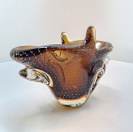 Centro de mesa em vidro Murano artístico âmbar com inclusões de bolhas. Peça escultórica em vidro artístico soprado, de forma orgânica e movimento fluido, com duas pontas ascendentes e base espessa translúcida. Apresenta tonalidade âmbar mel, com o uso da técnica bullicante  pequenas bolhas distribuídas de forma regular no interior do vidro  e o recurso sommerso, no qual camadas translúcidas são sobrepostas criando profundidade e brilho intensos. O trabalho revela refinamento técnico e domínio no manuseio do vidro quente, resultando em uma forma que equilibra força e leveza visual: 18 x 27 x 11 centímetros.