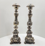 Majestoso par de tocheiros sacros em metal banhado a prata, de composição vertical e fuste balaústre repuxado e cinzelado em relevo com figuras sagradas representando Jesus Cristo, Nossa Senhora e São José. As bases tripartidas repousam sobre pés em volutas elegantemente modeladas, ornadas com folhagens e concheados. O conjunto apresenta exuberante ornamentação inspirada na arte sacra barroco-rococó portuguesa. Peças de caráter devocional e forte presença cenográfica, adequadas para altares, capelas ou oratórios domésticos. Dimensões: 53 x 17 centímetros.